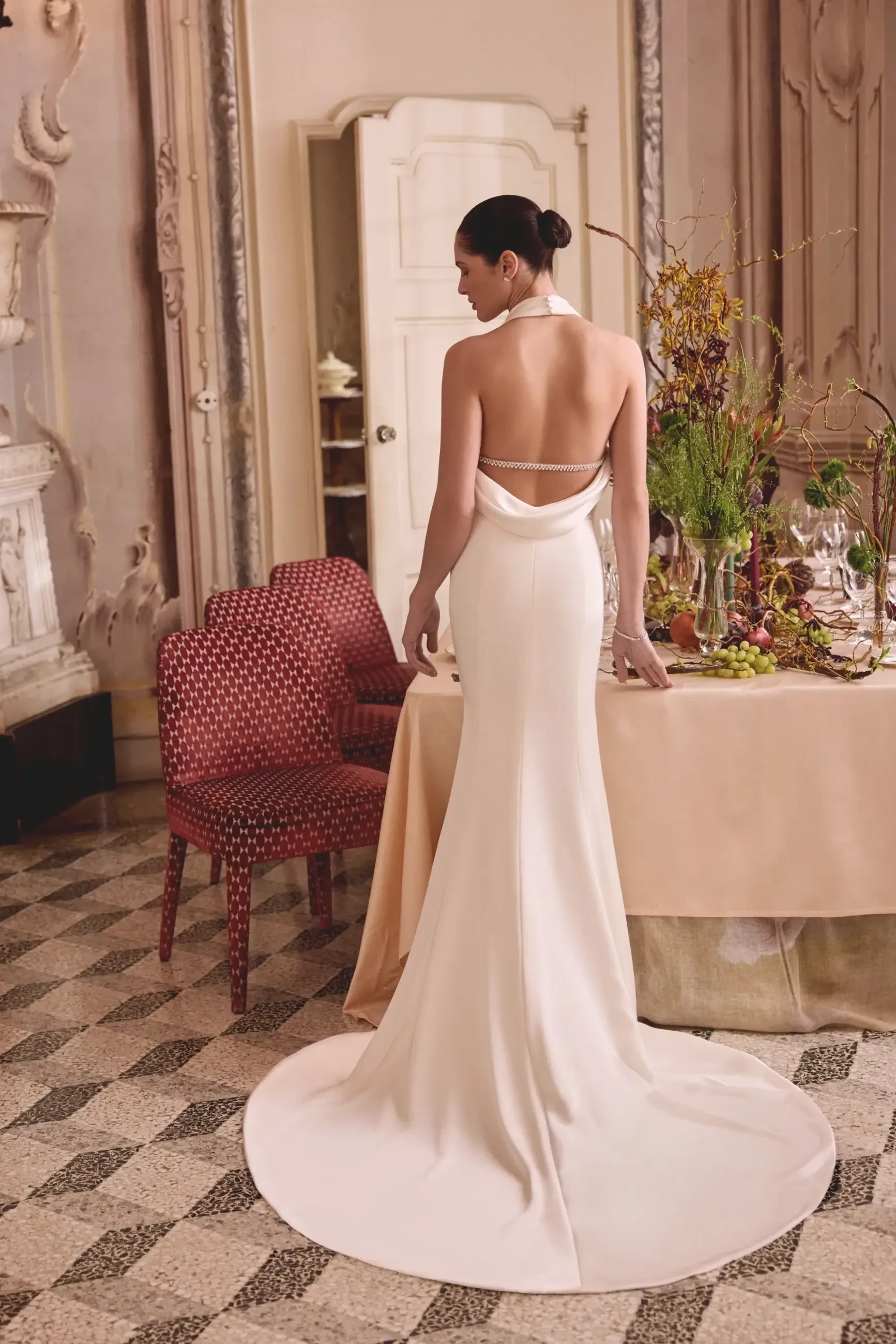 A woman in a sleek, white gown with an open back stands gracefully, facing away from the viewer. She is inside an elegant room with ornate decor and a dining table adorned with flowers and fine dinnerware.