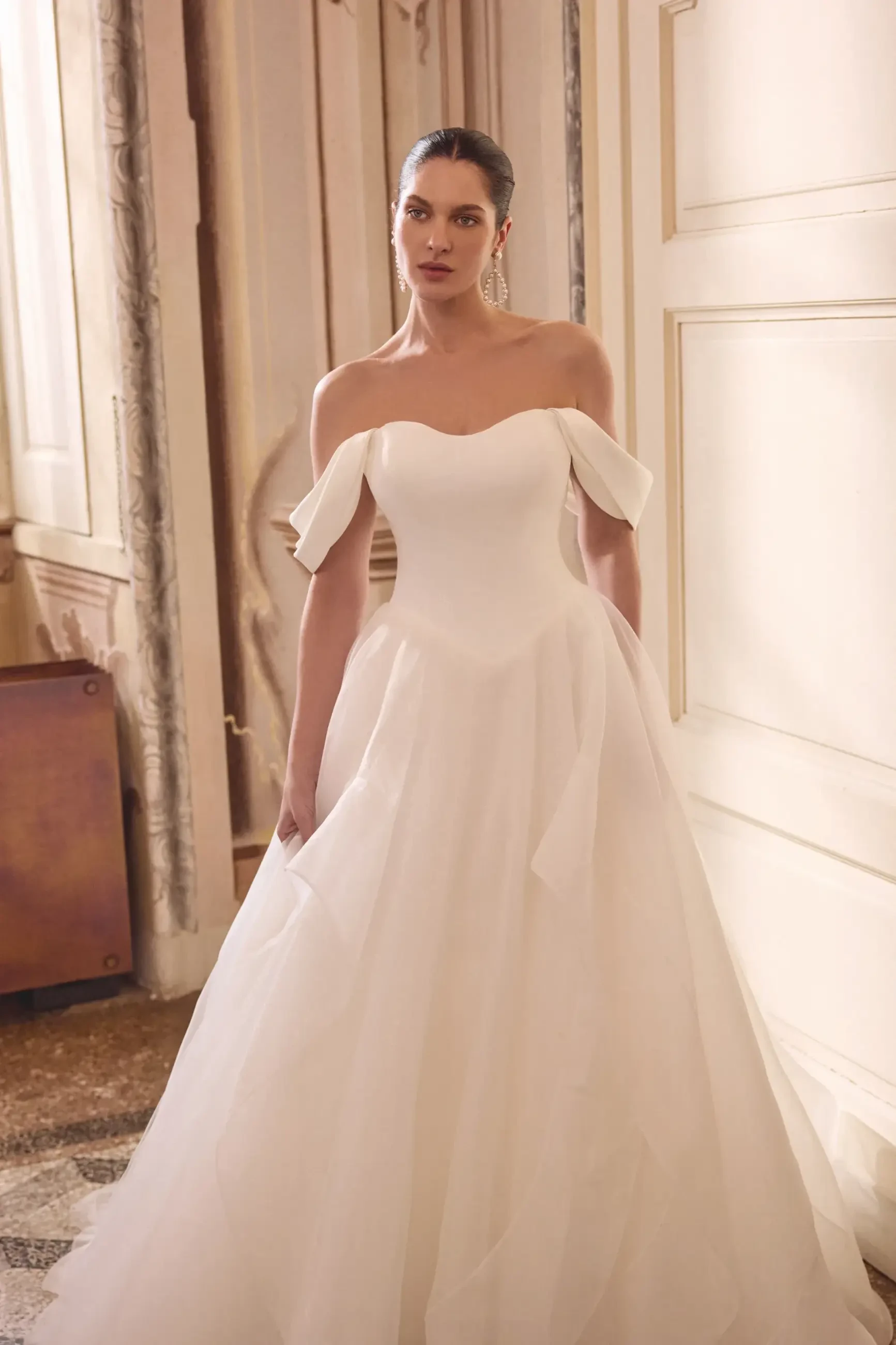 A woman stands in an elegant, sunlit room wearing an off-the-shoulder white wedding gown with delicate ruffles, conveying a serene and graceful mood.