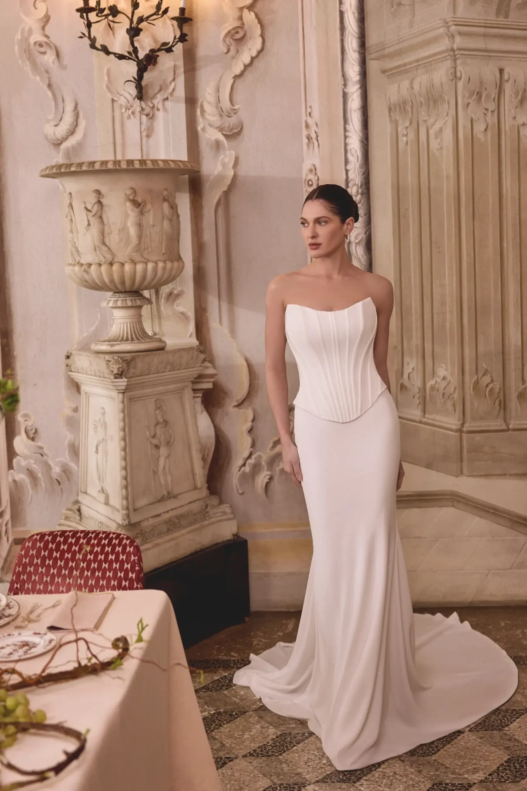 A woman wearing a white, strapless gown with a fitted bodice and flowing skirt, standing elegantly in a decorative room with ornate details.