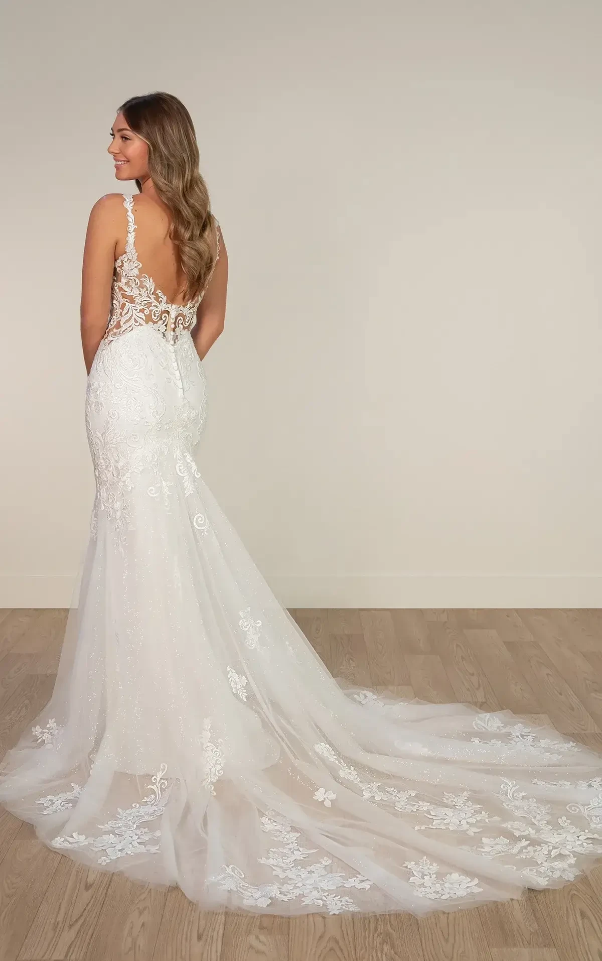 A woman is wearing a detailed lace wedding gown with an open back and long train. She is standing on a wooden floor, looking over her shoulder with a soft smile.