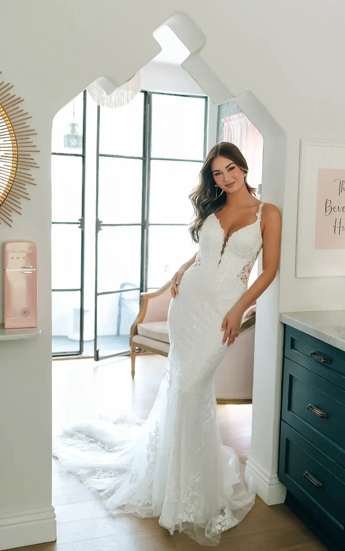 A woman in a lacy white wedding dress stands in a bright, elegant room by a large window. She smiles softly, conveying happiness and elegance.