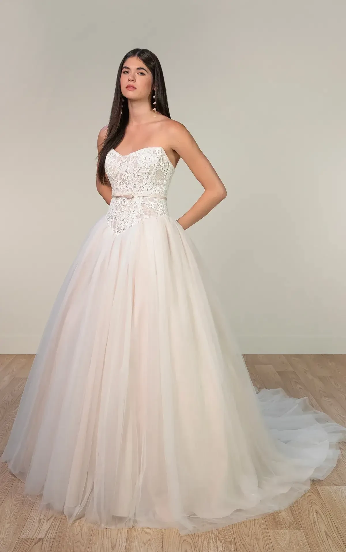 A model is wearing a strapless white gown with a fitted bodice and a voluminous tulle skirt. The dress has decorative embellishments on the bodice and flows elegantly to the floor.