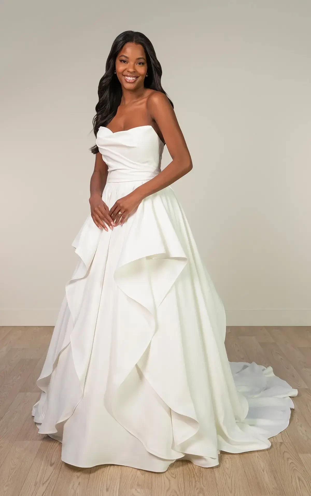 A woman wearing a strapless white wedding gown with layered ruffles, standing and smiling in a neutral-colored room.