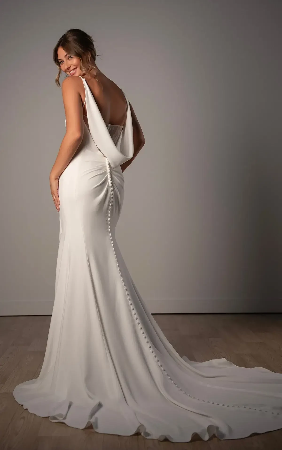 A woman in a white bridal gown with a draped back and buttons running down the skirt, standing sideways and looking over her shoulder.
