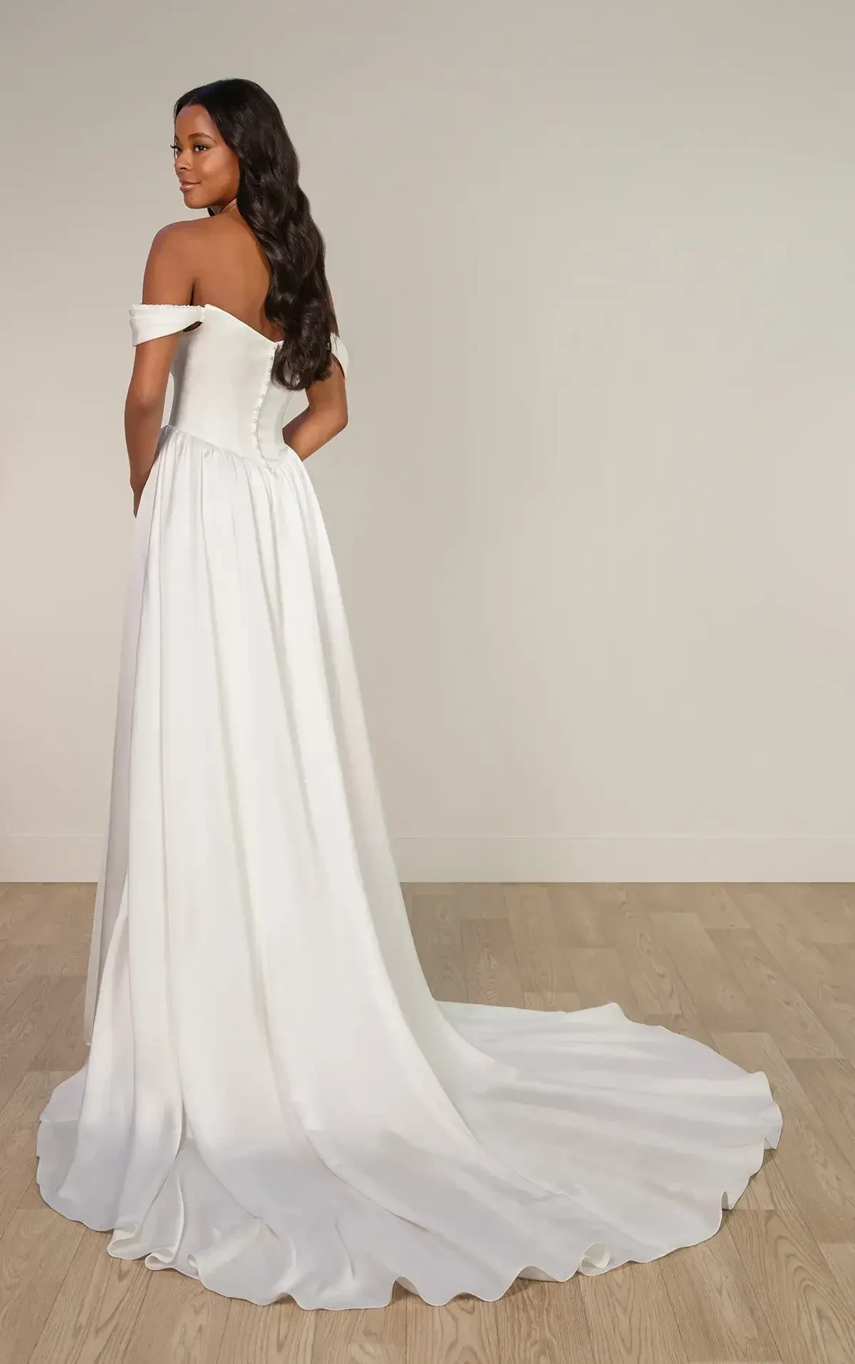 Elegant bridal gown with an off-shoulder design and a long, flowing train. The woman stands on a wooden floor, exuding grace and sophistication.