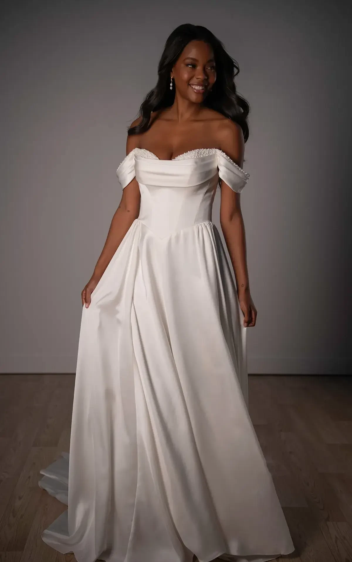 A woman in an elegant, off-shoulder white wedding dress smiles warmly. The gown features a fitted bodice and flowing skirt, exuding a joyful, graceful ambiance.